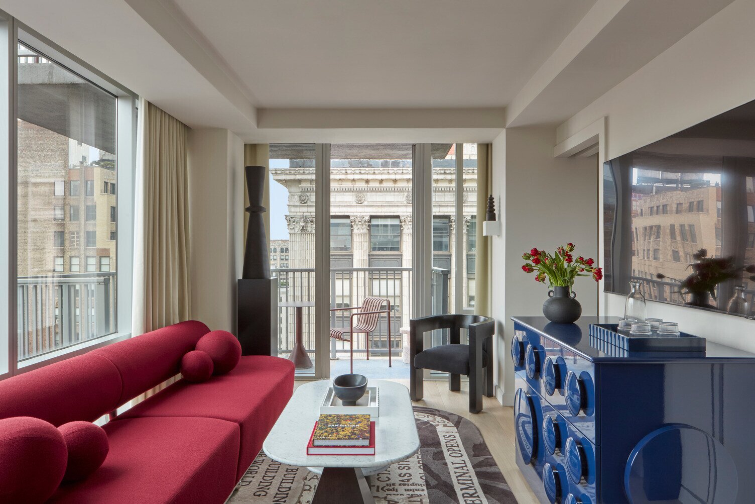 Studio Balcony Suite with bold red curved sofa, round accent cushions, large windows, blue glossy cabinet, panoramic city views, abstract artwork, balcony, and marble coffee table with decorative fruit bowl and vases.