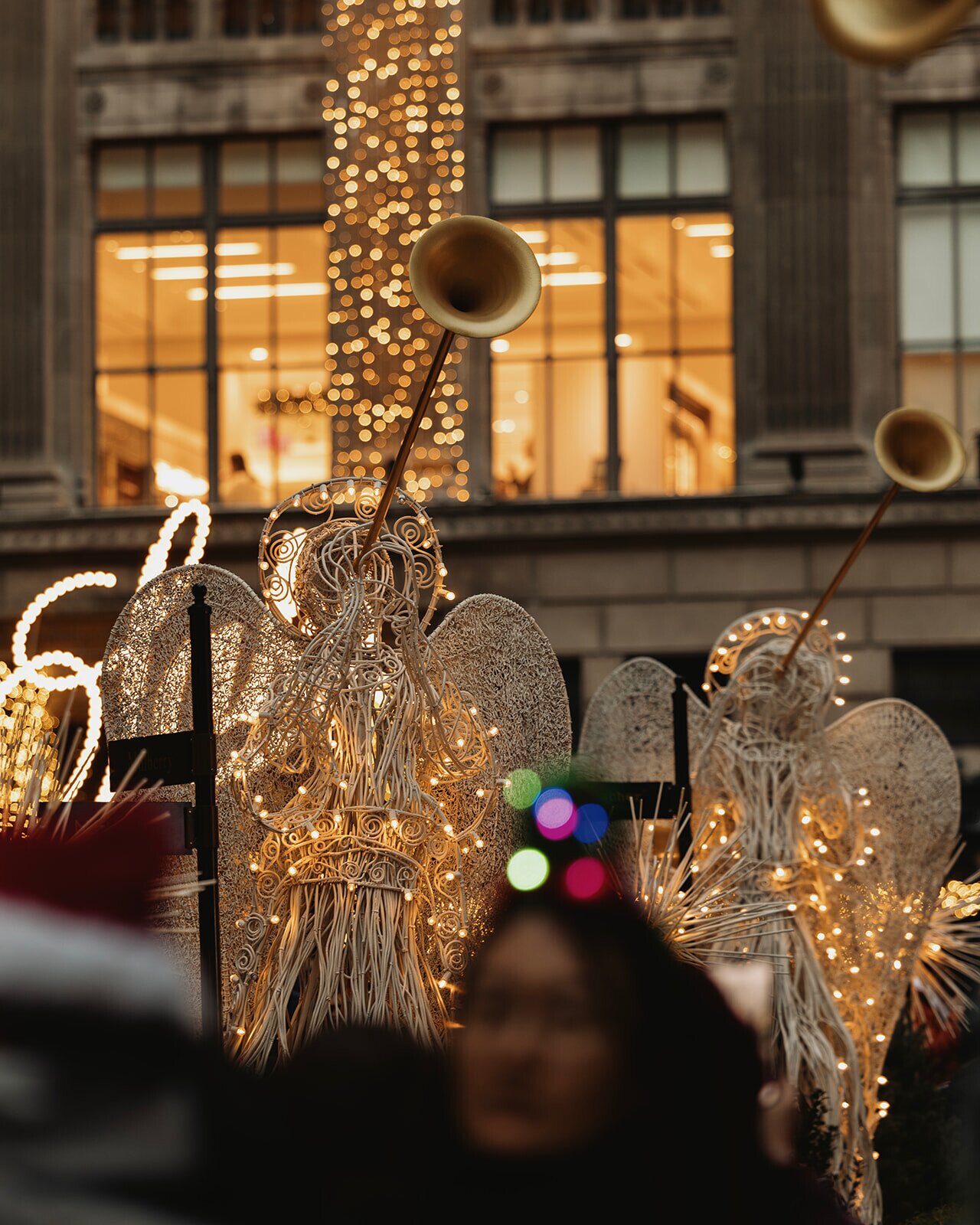 Lit angel trumpet decorations along Rockefeller Center’s holiday walkway, with glowing Christmas lights and New York City storefronts in the background.